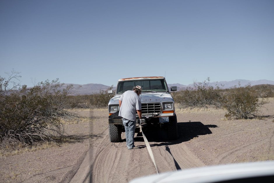 A man at the front of a stranded pickup truck on a desert road, with a tow strap stretched back to the recovery vehicle